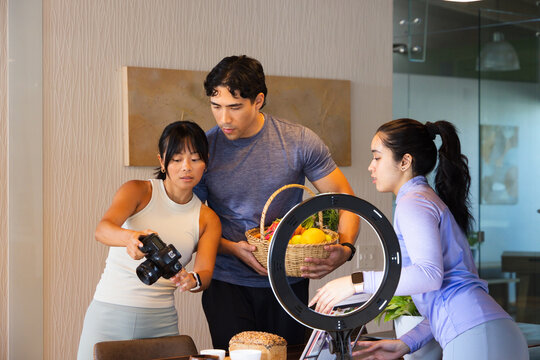 Diverse coworkers reviewing DSLR at studio, adjusting ring light, holding wicker basket with loaf