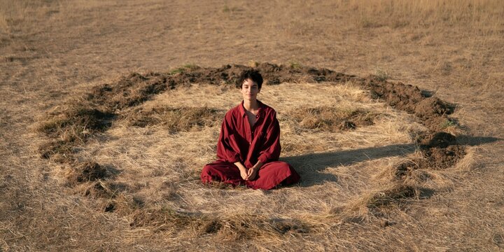 Meditation monk in red robe seated inside a circular earth trench in dry field for wellness background and contemplative banner design