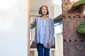 Naklejka premium Senior Asian woman standing in doorway at home wearing gray cardigan and jeans, holding carved door
