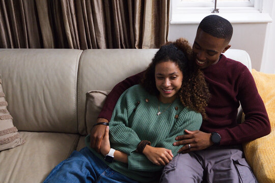 Diverse couple sitting cuddling on light beige leather sofa at home with watches visible