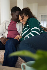 Obraz premium African American couple sitting on couch at home, leaning together, green knit, blurred plant, mug