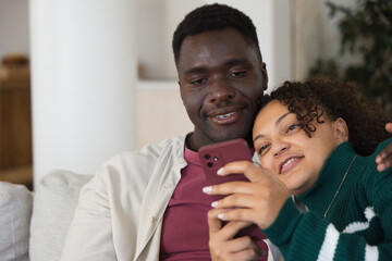 Obraz premium African American couple sitting on light-colored sofa at home, holding red smartphone and smiling