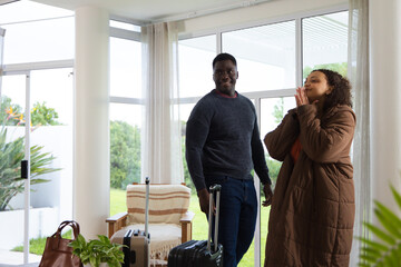 Obraz premium African American couple standing in living room by sliding doors with luggage wearing brown coat