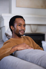Obraz premium Adult African male sitting on gray sofa wearing brown quilted sweater near white mantel and artwork