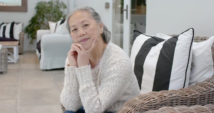 Senior Chinese woman resting chin, rubbing neck, settling in wicker chair, relaxing in sunroom