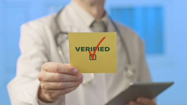 Doctor in a white lab coat and stethoscope holding a yellow note with the word verified and a red checkmark, symbolizing professional certification and approval in healthcare
