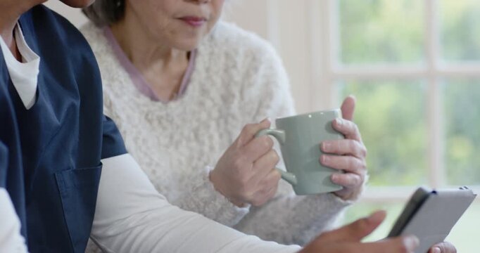 Nurse in scrubs tapping tablet, explaining meds while Senior Chinese woman holding mug by window