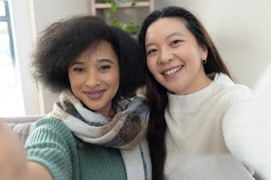 Diverse female friends taking selfie on couch with smartphone wearing green knit sweater and scarf