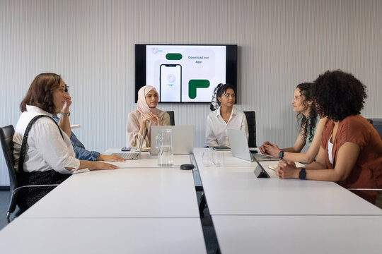 Diverse female colleagues discussing app mockup on wall monitor at conference table with laptops