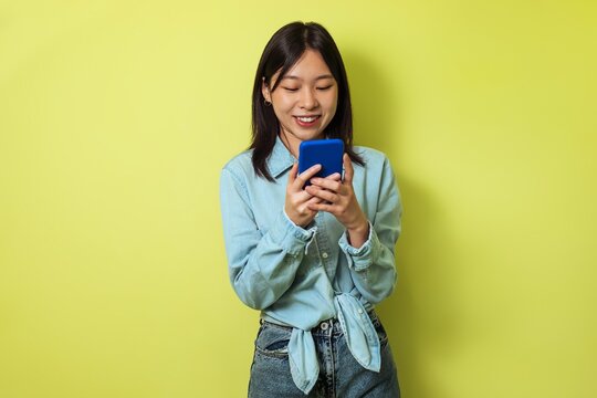 Smiling Young Asian Woman Holding Smartphone With Blue Case, Texting While Standing Against Bright Yellow Background In Modern Studio. Showing Joy And Engagement With Mobile Technology.