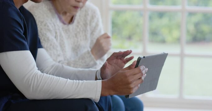 Diverse male carer guiding senior Asian woman on couch, tapping gray-cased tablet after prompt