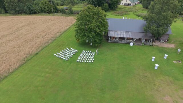 Aerial view of outdoor wedding setup with white chairs arranged in rows on green grass near a rustic barn surrounded by trees and fields in a serene countryside setting