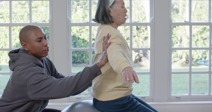 Senior Chinese woman balancing on ball in home while African helper placing hands guiding mobility