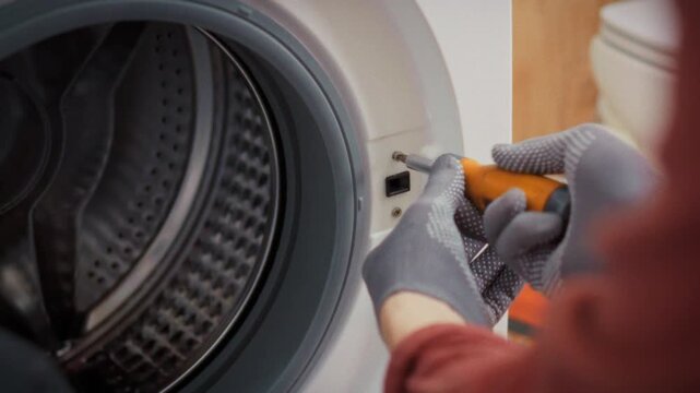 A close-up of a screwdriver in a technician's hands unscrewing a bolt from a washing machine body in a bathroom with a toolbox in the background. Concept of domestic appliance repair
