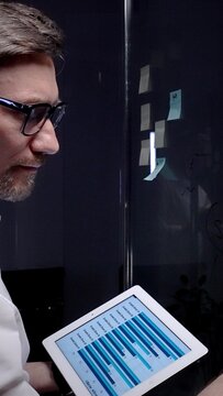 Business professional analyzing data on a tablet and planning strategy with adhesive notes on a glass board, brainstorming and organizing ideas for project development in a modern office