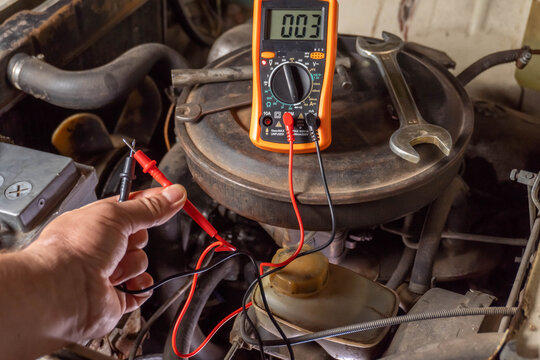 A hand holds multimeter probes as readings are taken from an engine component, showcasing routine car maintenance inside a garage. Tools and wires are neatly arranged, highlighting precision