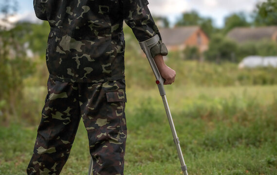 A soldier in camouflage stands on crutches, looking out over a serene settlement. The scene depicts the moment of returning home after being wounded