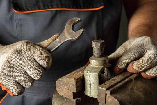 A focused individual uses a wrench on a metal clamp within a bustling workshop. Gloves protect hands while the workbench is cluttered with tools and equipment
