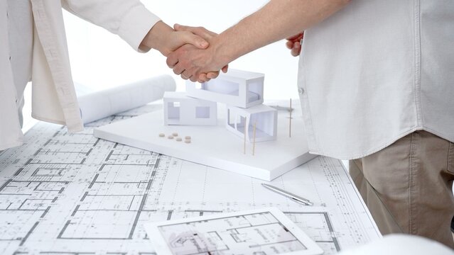 Business partners shaking hands over architectural blueprints and a building model, finalizing a real estate project agreement and demonstrating successful collaboration