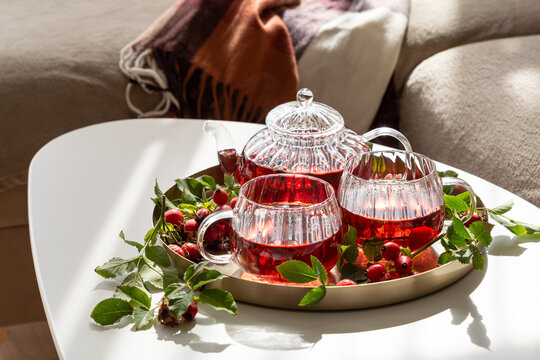 Rosehip tea in glass teapot and cups with fresh berries and green leaves on tray