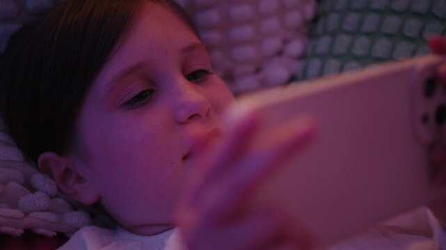 Close-up of a young girl lying in bed under soft ambient light while looking at a smartphone screen late at night.