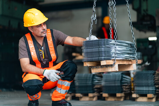 Construction worker in safety gear handling steel wire bundle with overhead chain hoist in industrial warehouse, focused laborer preparing heavy material for transport and manufacturing work