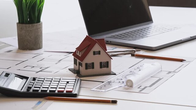 Tiny house model sits on desk beside laptop. Blueprints, pencils, calculator, and green plant complete scene. No people, so no emotions to describe. Perfect for real estate, construction