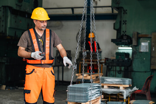 Industrial worker wearing safety gear lifts stacked metal sheets with chain hoist inside dark workshop, showing careful handling and focused work