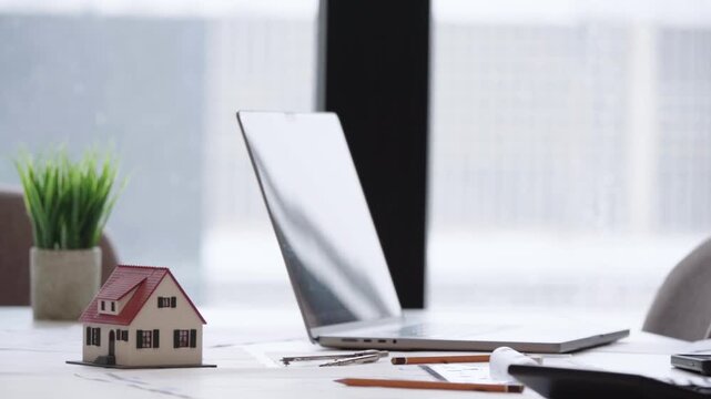 Mini house sits beside laptop on desk. Pencil and papers lie nearby. Green plant adds subtle life to workspace. Focused mood suggests real estate or planning. Perfect for finance, property