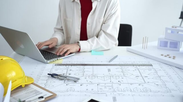 Architect working on new residential project using laptop, reviewing blueprints and drafting floor plans for modern building design with a 3d model on office desk