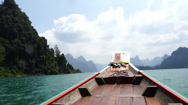Wooden boat glides through turquoise waters. Lush green hills rise on either side. Sky holds soft clouds above. Calm waters reflect distant peaks. Serene journey through quiet jungle river