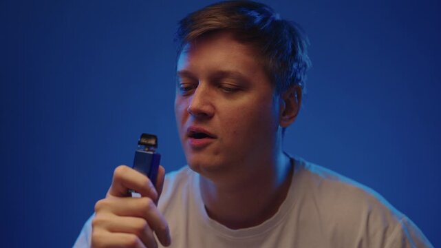 Portrait of casual young adult man smoking vaping device in dark studio setting, puffing on ecigarette and blowing out thick cloud of vapor with relaxed expression on face, close-up, slow motion.
