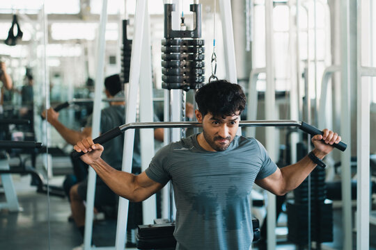 Young man performing lat pulldown exercise on gym weight machine, focused fitness training with strength equipment in bright modern workout facility
