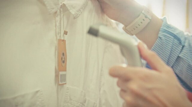 A store manager runs a red laser scanner over a barcode on a clothing label carefully registering the item in the store database against a background of hanging garments