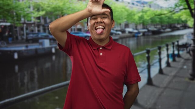 Man wearing red polo sticks out tongue and forms l with hand to forehead on street by canal in amsterdam; playful mischief.