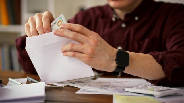 At his work desk, a man's hands carefully sort crisp banknotes into individual envelopes, surrounded by paperwork. The routine captures the classic office practice of issuing cash wages to employees