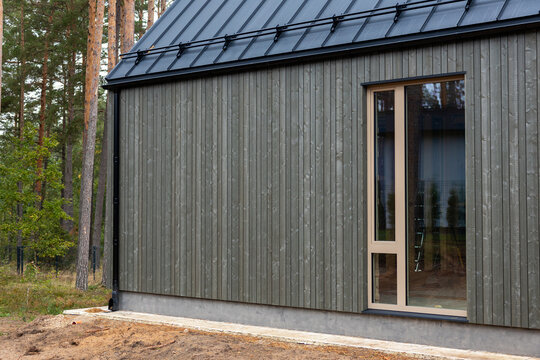 Modern wooden house facade with vertical timber planks and metal roof stands beside pine woods during daylight, showing exterior design details and tall window.