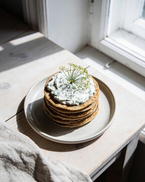 Tall stack of buckwheat blini with herb cream cheese topped with dill flower by a bright window.