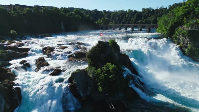 Aerial slow orbital shot of the central rocky island with a waving Swiss flag at the Rhine Falls in Schaffhausen, featuring wide river rapids.