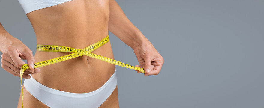 Focused Woman Measuring Waist With Tape Measure While Standing In A Neutral Background, Taking Control Of Her Body Measurements And Progress Towards Weight Loss Goals. Weight Loss Concept.