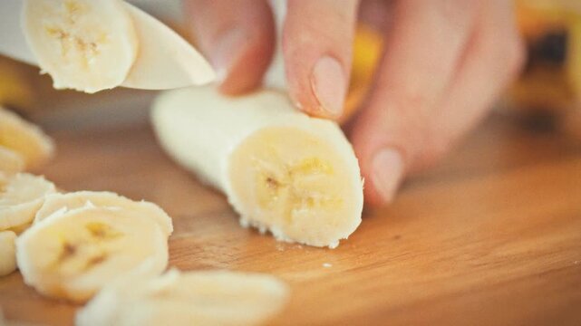 In bright summer light, man's hands slice a ripe banana on a wooden board, the crisp cut revealing fresh fruit. The simple kitchen moment captures the joy of preparing a tropical snack on a sunny day