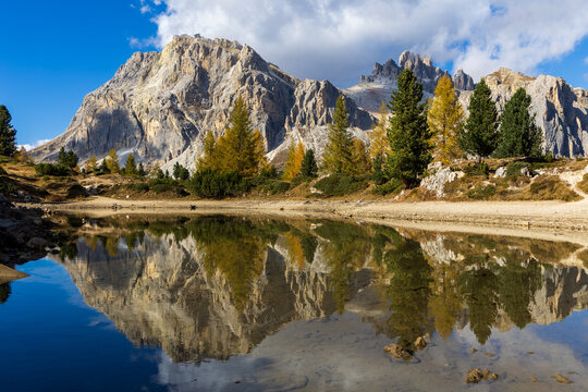 An autumn landscape featuring golden larches and surrounding mountains is reflected in the calm waters of Lago di Limedes near Passo Pordoi in the Dolomites.