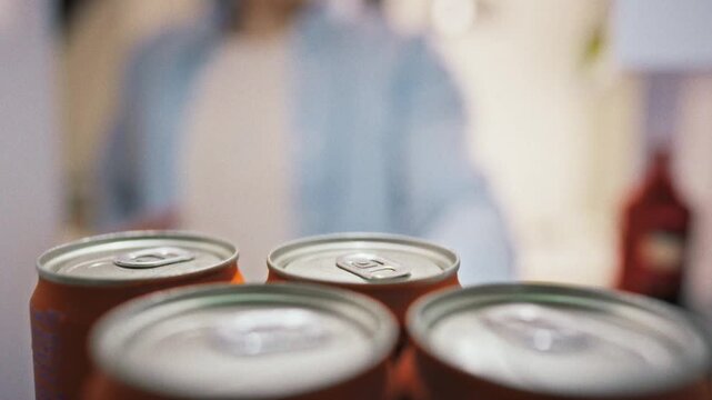 A man in casual home clothes lines up tin cans of carbonated drinks on a refrigerator shelf, his hand steady as he arranges each can inside the kitchen fridge, a routine food-storage task