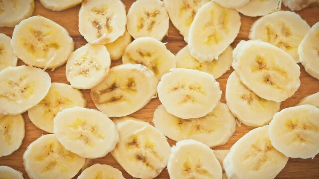 Thinly sliced banana pieces are randomly scattered across a wooden board, illuminated by warm sunlight. Concept for fresh fruit for breakfast and cooking