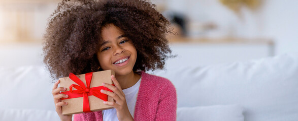 Naklejka premium Happy Black Girl Smiling While Holding Gift Box With Red Ribbon, Displaying Excitement At Home Setting. Joyful Young Child Celebrating Special Occasion Or Birthday With Present.