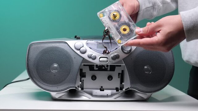Person's hands holding a broken clear cassette tape with its magnetic tape pulled out and tangled above a silver vintage boombox with an open tape deck on a white surface