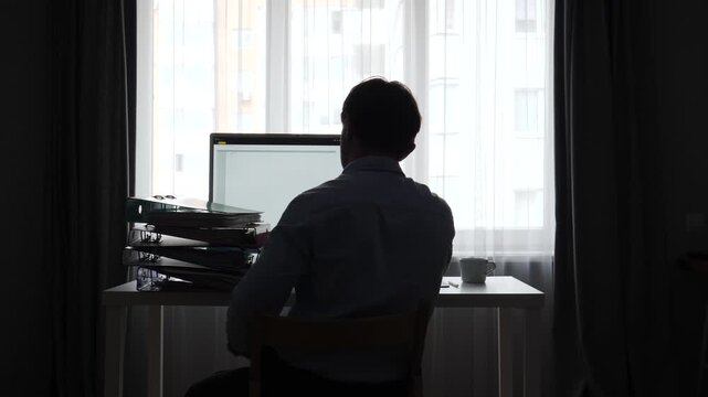 Business person in silhouette sitting at a desk, looking at a computer screen, with a tall stack of folders and documents on the side, representing long hours and heavy workload