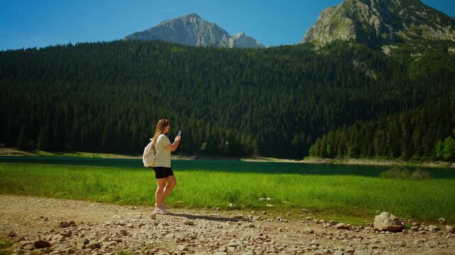 Young woman walking along tranquil mountain lake shore, capturing scenic forest landscape and towering rocky peaks under clear blue summer sky on hiking adventure