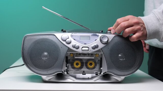 Hand pressing buttons on a silver retro boombox with a cassette tape visible, listening to music or tuning a radio station on a white table with a teal background