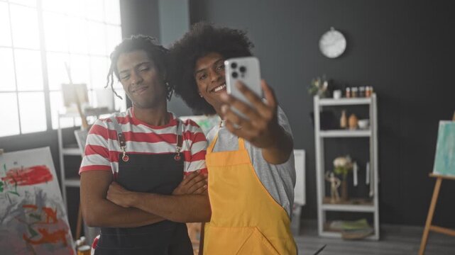 Man painter holding smartphone for selfie, arms crossed and wearing aprons beside easel and canvas in studio; playful collaboration.